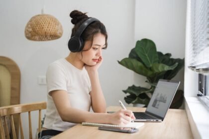 A woman sitting at her kitchen table, wearing headphones and working on a laptop.